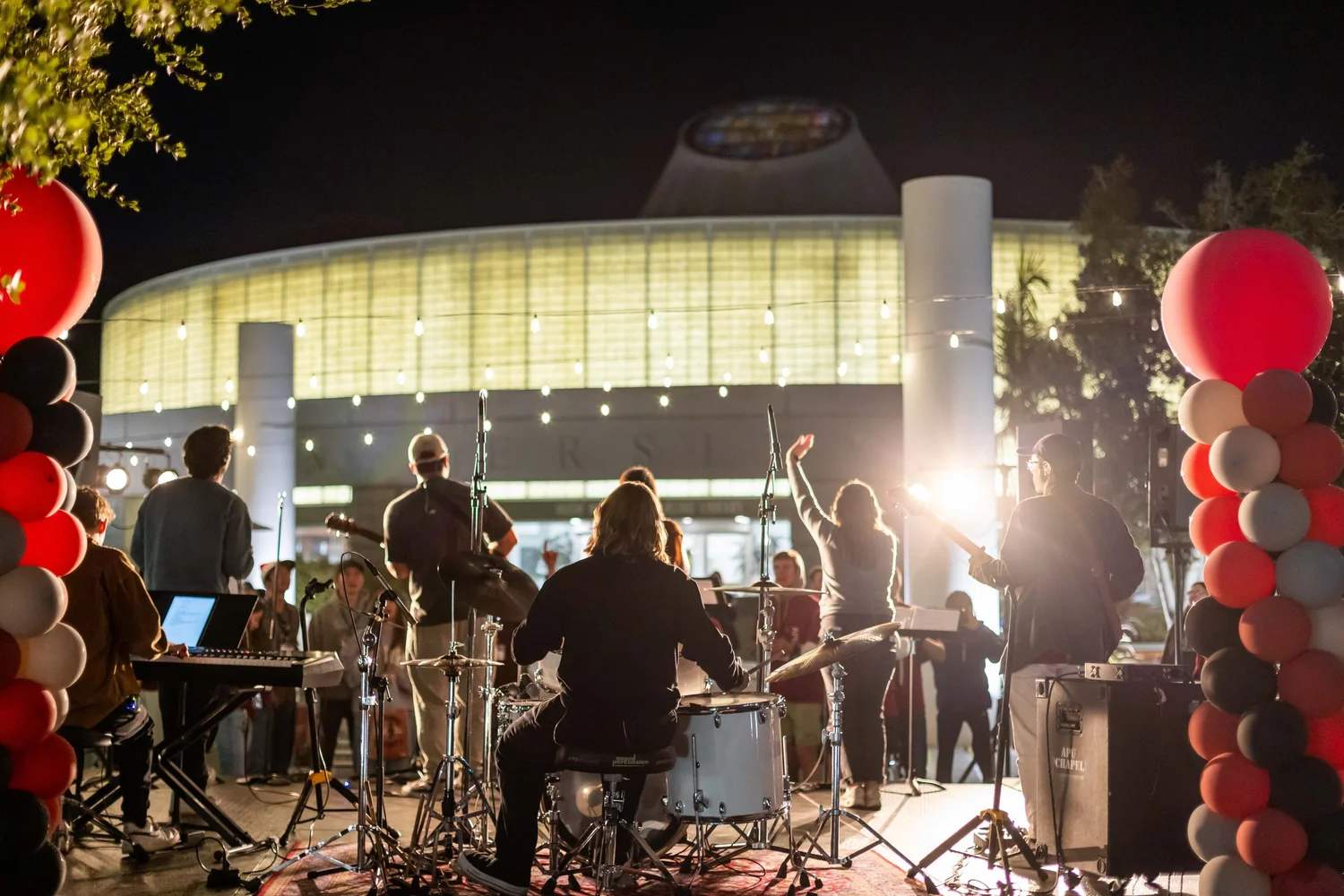 Live band performing for a crowd at a nighttime campus event framed by red and white balloons