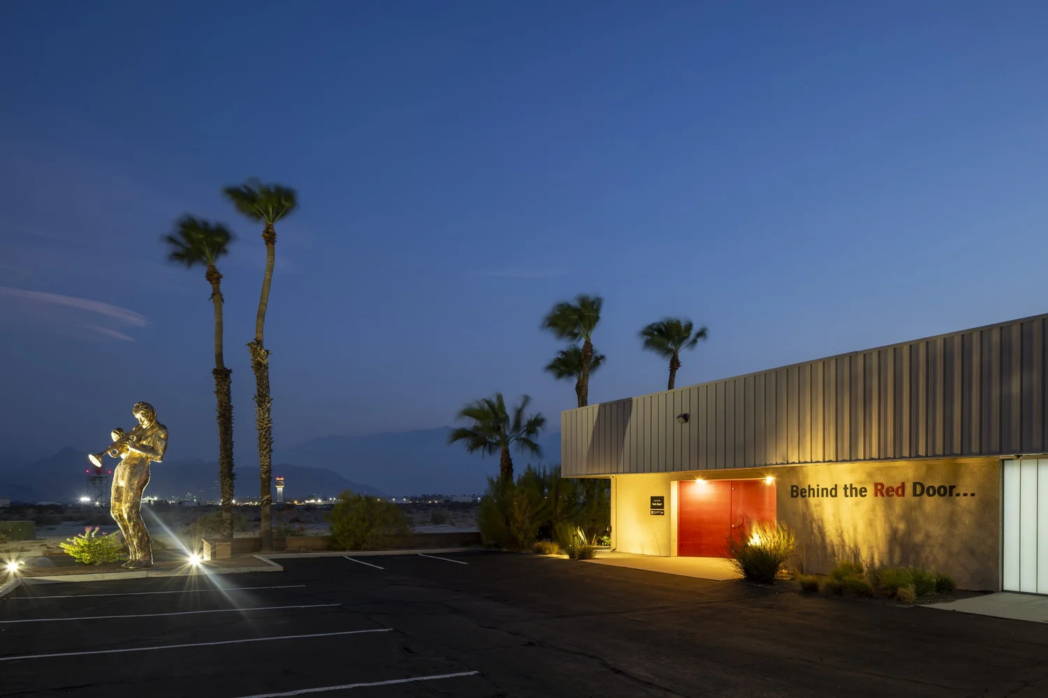 Herb Alpert Gallery of Art in Palm Springs at dusk, with palm trees, a gold figure sculpture, and a glowing red door