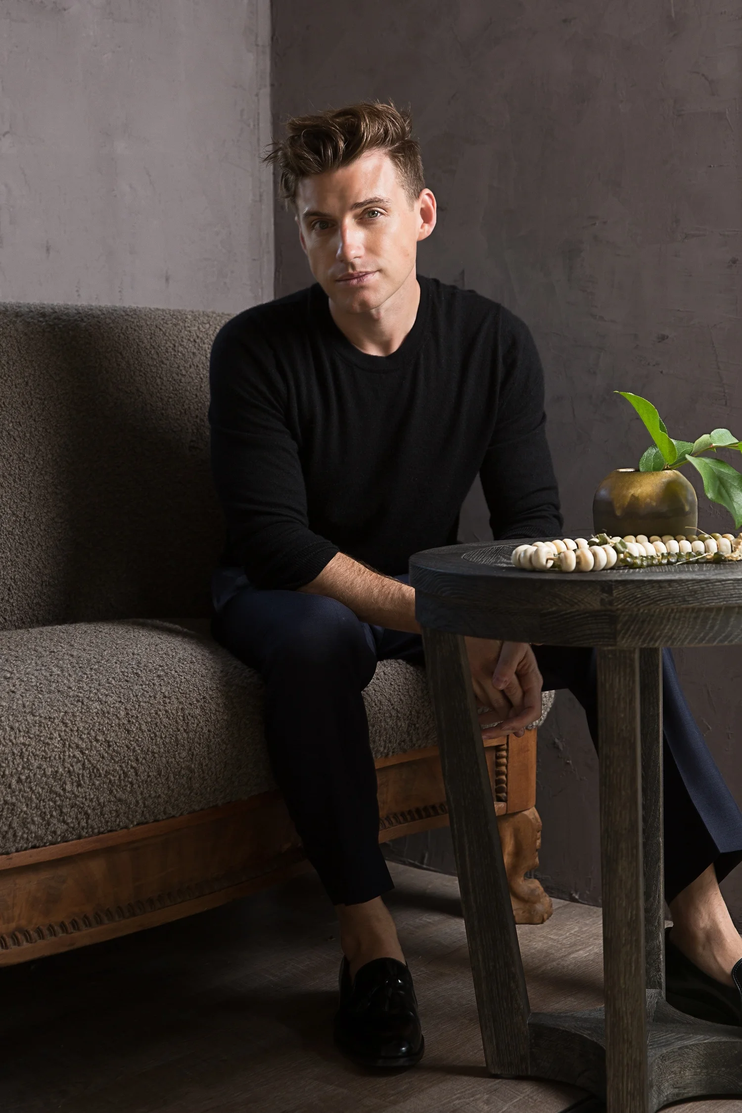 Portrait of designer Jeremiah Brent in black, seated at a small table against a concrete wall